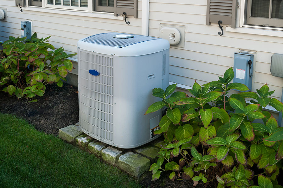 Residential air conditioning system like Ruud, Amana, and Heil attached to a cream-colored home and surrounded by large green bushes in the Metro East area.