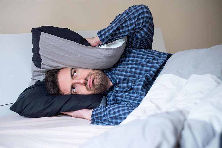 A man in his pajamas lying down on a bed in his Metro East home holding a pillow over his ears to drown out a noisy furnace that needs to be upgraded.