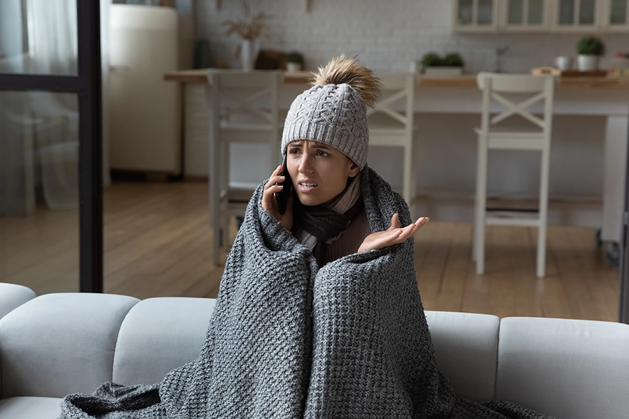 An unhappy female homeowner wrapped in a blanket and winter gear because her furnace is not operating properly in her home in the Metro East Area of IL—she is confused why it is emitting cold air instead of warm air.